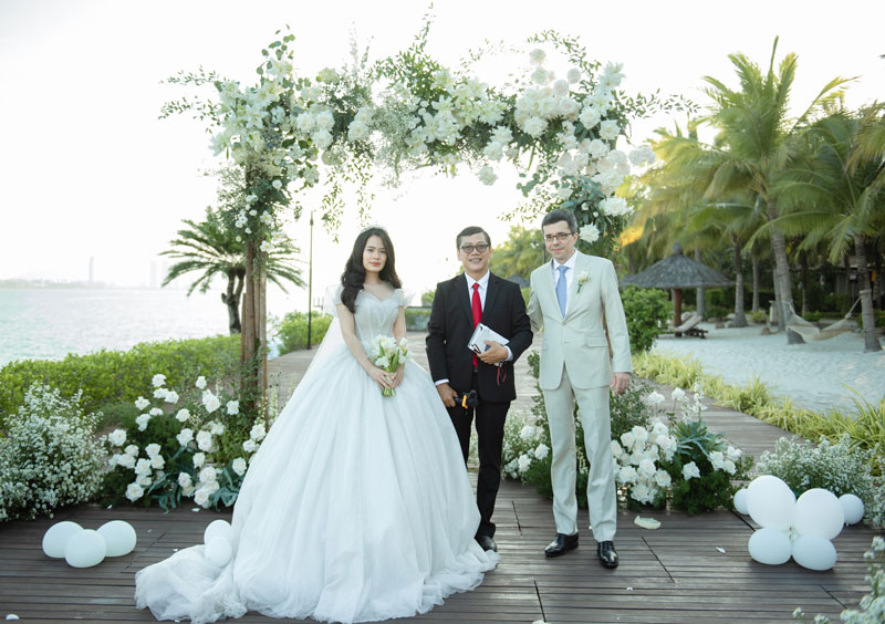 Cô dâu váy trắng, chú rể vest kem và chủ lễ dưới cổng hoa trắng tại Amiana Resort Cô dâu váy trắng, chú rể vest kem và chủ lễ dưới cổng hoa trắng tại Amiana Resort