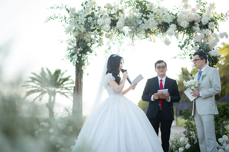 Cô dâu cầm micro phát biểu dưới cổng hoa trắng, nắng chiều hắt từ phía biển Cô dâu cầm micro phát biểu dưới cổng hoa trắng, nắng chiều hắt từ phía biển