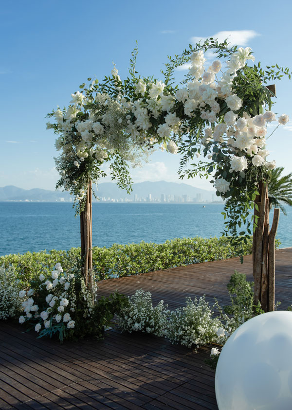 Trang Trí Tiệc Cưới Ngoài Trời Nha Trang: Amiana, Alibu Cổng vòm gỗ thô phủ hoa trắng trên sàn gỗ Amiana, phía sau là vịnh và thành phố Nha Trang