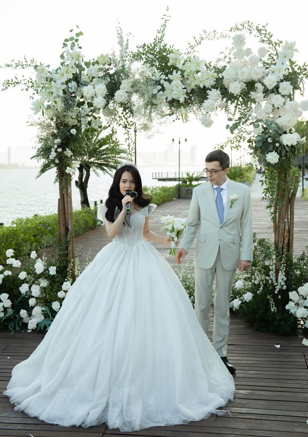 Bride speaking into a microphone under the white flower arch beside the groom in a cream suit Bride speaking into a microphone under the white flower arch beside the groom in a cream suit