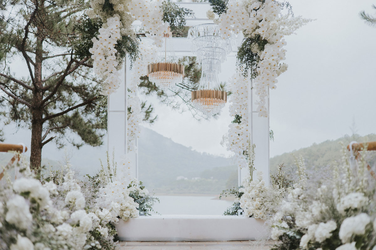 Cổng trụ trắng phủ lan hồ điệp trắng rủ, đèn chùm pha lê, phía sau là hồ và đồi thông Cổng trụ trắng phủ lan hồ điệp trắng rủ, đèn chùm pha lê, phía sau là hồ và đồi thông