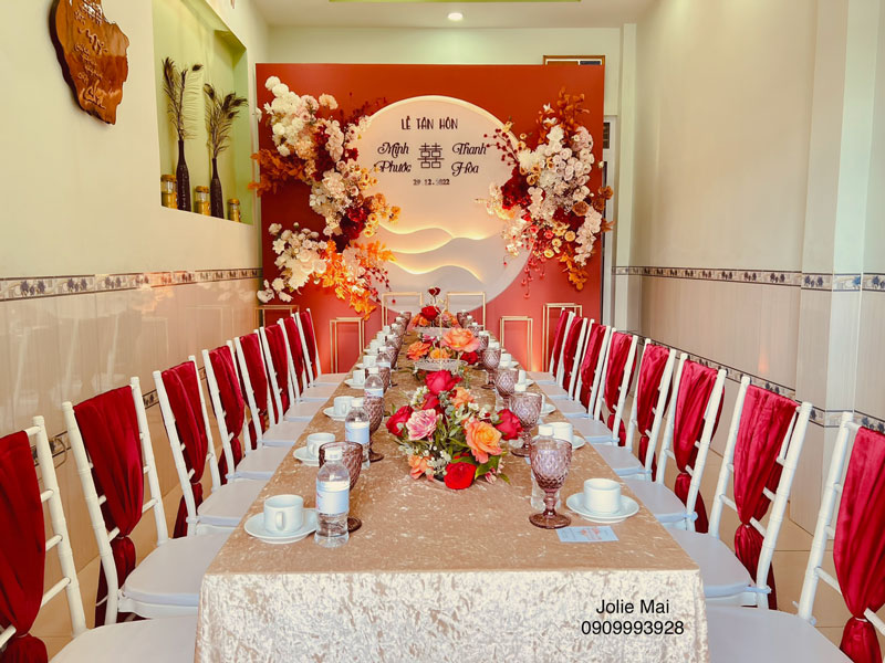 Gia Tiên Đỏ Đô: Phông Cánh Sóng, Nhung Đỏ Mận, Hoa Kem Phông cam đỏ vòng tròn sáng, bàn dài khăn ren kem, ghế trắng buộc nhung đỏ trong phòng hẹp