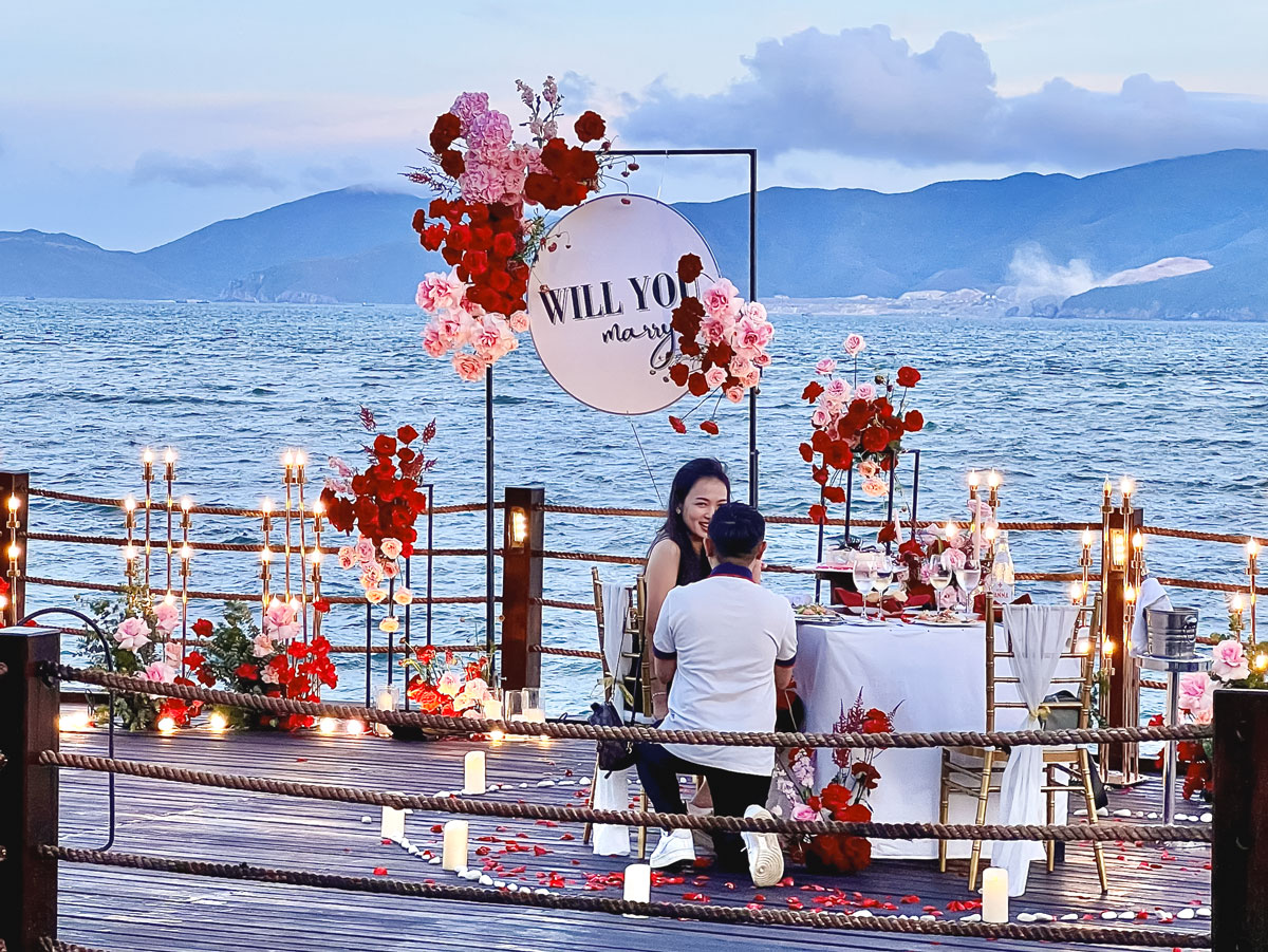 Cầu gỗ ven biển tại Amiana Resort với bảng tròn Will You Marry và bàn ăn hai người Cầu gỗ ven biển tại Amiana Resort với bảng tròn Will You Marry và bàn ăn hai người