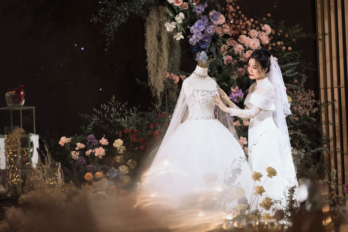Cô dâu chỉnh váy trên ma-nơ-canh đội vương miện, nền tối điểm sao với hồng đào, cẩm tú cầu tím và đèn dây Cô dâu chỉnh váy trên ma-nơ-canh đội vương miện, nền tối điểm sao với hồng đào, cẩm tú cầu tím và đèn dây