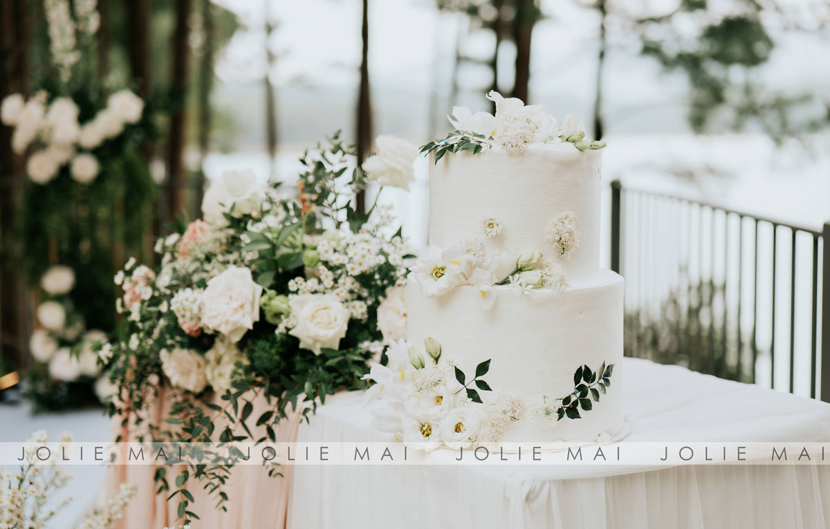 Two-tier white wedding cake ringed with white flowers, the lake behind the railing Two-tier white wedding cake ringed with white flowers, the lake behind the railing