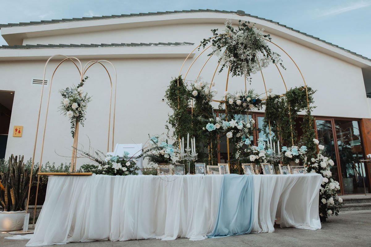 Wedding in Cam Ranh: Stunning Beach Vows and Resort Lawns Gallery table with framed photographs under white and pale blue fabric, beside gold arch frames with flowers at Mövenpick Resort Cam Ranh