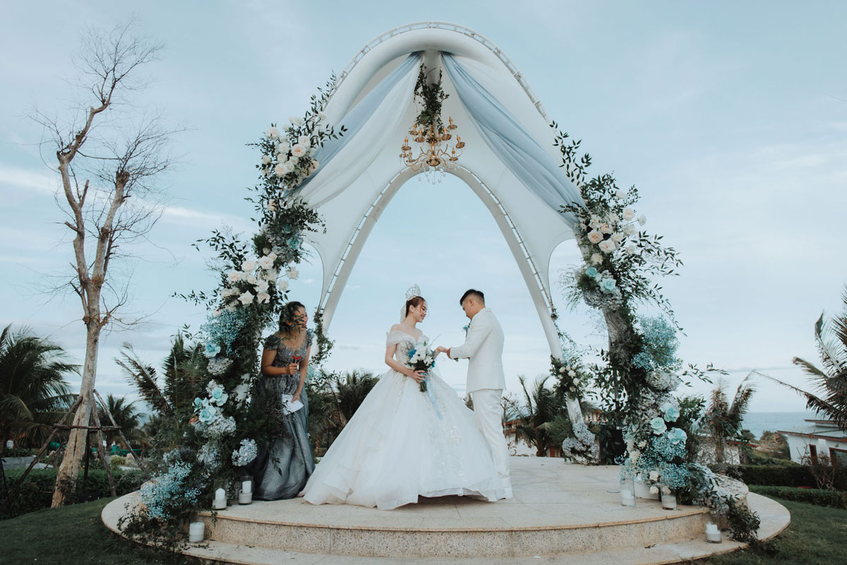 Trang Trí Tiệc Cưới Cam Ranh: Resort Bãi Dài Và Tiệc Tại Nhà Lễ thành hôn dưới vòm trắng phủ hoa xanh trắng trên bục đá, nhìn ra biển Bãi Dài