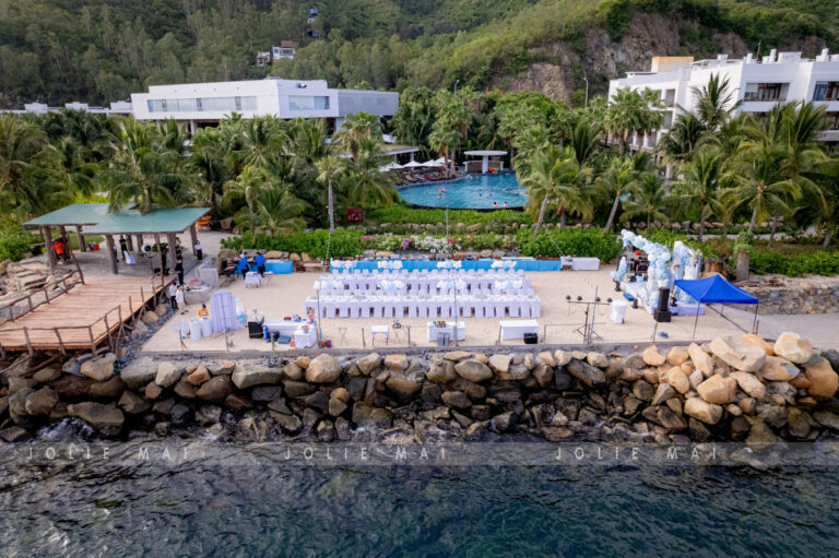 Trang Trí Sinh Nhật Nha Trang: Bãi Biển, Resort, Tại Nhà Dải cát có hai dãy bàn tiệc trắng và backdrop, phía sau là hồ bơi và khu resort trắng, nhìn xéo từ biển