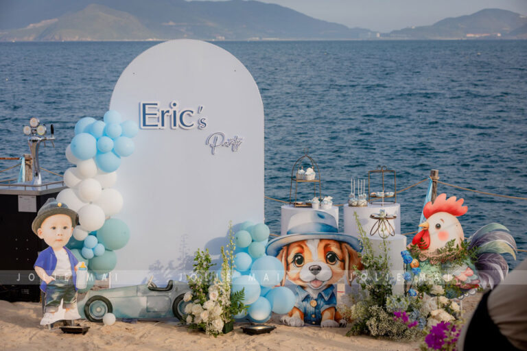 Trang Trí Sinh Nhật Nha Trang: Bãi Biển, Resort, Tại Nhà Trang trí sinh nhật Nha Trang: bảng vòm trắng chữ Eric's Party, standee bé trai và chú chó đội mũ cạnh bàn dessert bên biển