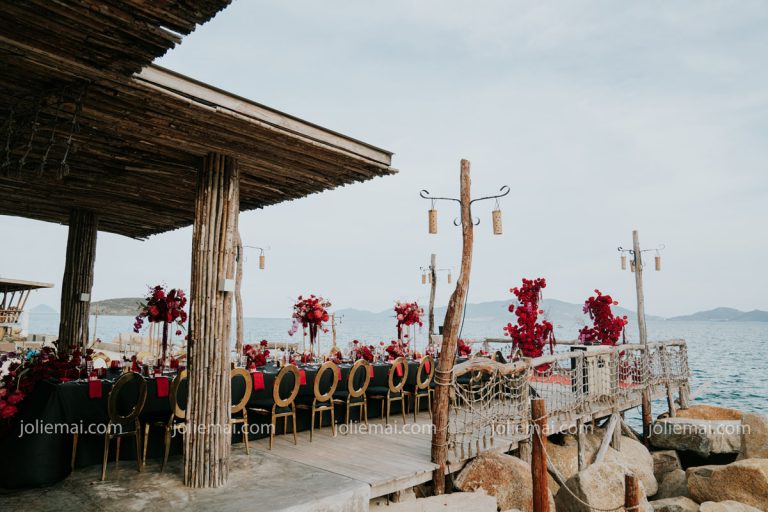 Dịch Vụ Cưới Nha Trang Một Đầu Mối: Gia Tiên, Tiệc, Mâm Quả Tiệc kỷ niệm ngày cưới trên sàn gỗ ven biển: bàn dài khăn đen, ghế vàng lưng tròn, cụm hoa đỏ cao dọc bàn