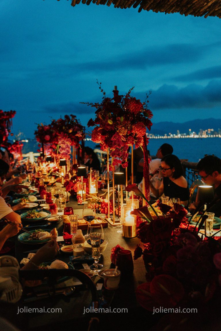 Trang Trí Tiệc Nha Trang: Sinh Nhật, Kỷ Niệm, Tất Niên Và Giáng Sinh Tiệc kỷ niệm buổi tối bên vịnh: nến, đèn bàn và hoa đỏ trên bàn dài, đèn thành phố phía xa