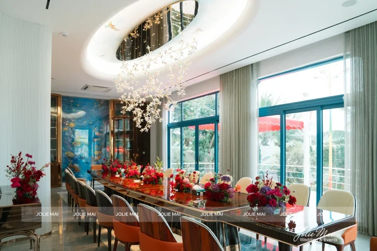 Trang Trí Gia Tiên Nha Trang: Hoa Lụa Từ 9,8 Triệu Bàn tiệc dài gỗ tối trong phòng ăn, lọ hoa đỏ đặt dọc bàn dưới đèn chùm