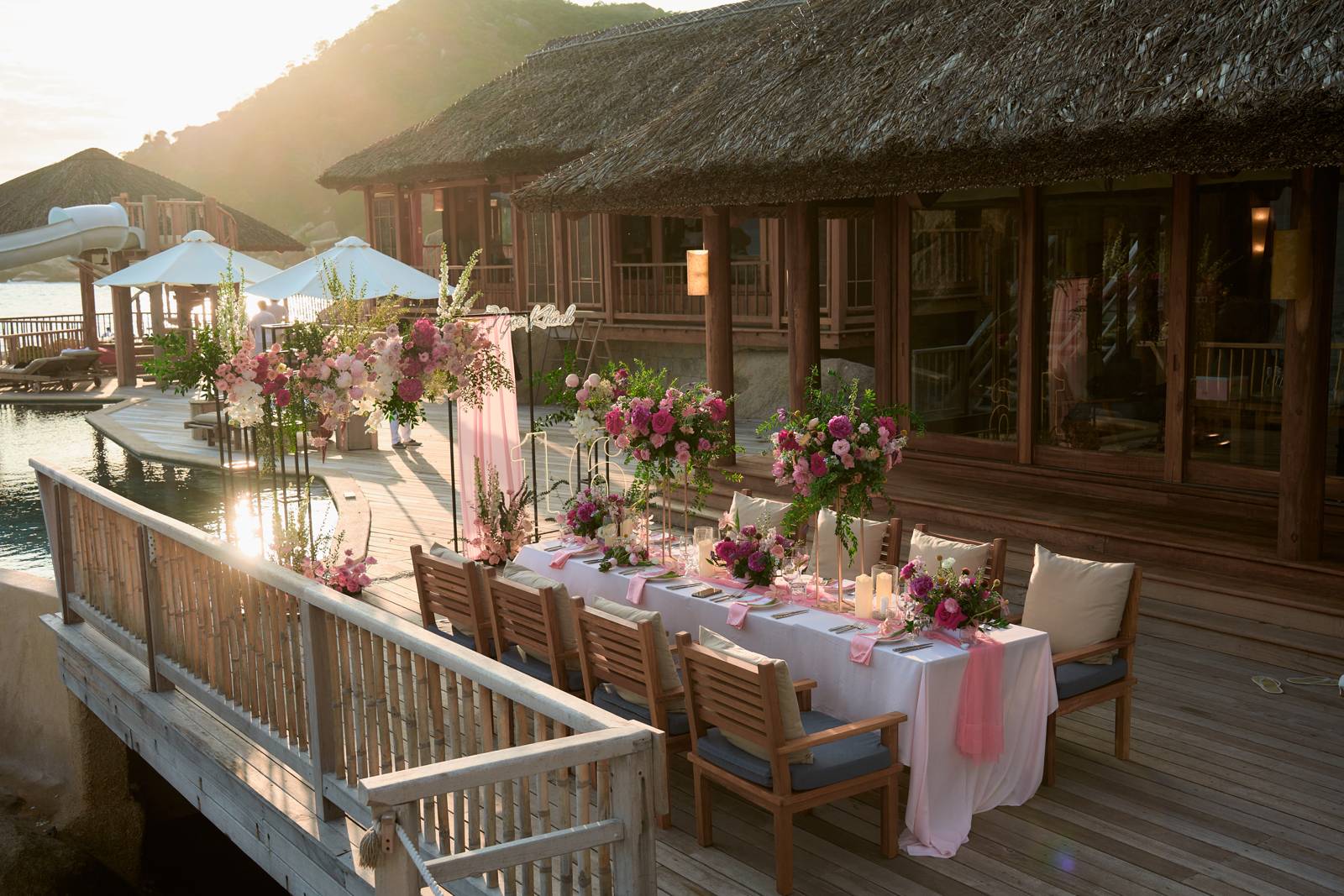 Bàn tiệc dài khăn trắng nơ hồng trên sàn gỗ cạnh hồ bơi vách đá, mái lá ở phía sau tại Six Senses Ninh Vân Bay Bàn tiệc dài khăn trắng nơ hồng trên sàn gỗ cạnh hồ bơi vách đá, mái lá ở phía sau tại Six Senses Ninh Vân Bay