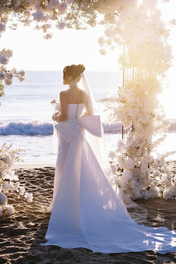 Cô dâu quay lưng đứng trước cổng hoa trắng trên bãi cát, sóng biển và ánh sáng cuối ngày phía sau Cô dâu quay lưng đứng trước cổng hoa trắng trên bãi cát, sóng biển và ánh sáng cuối ngày phía sau