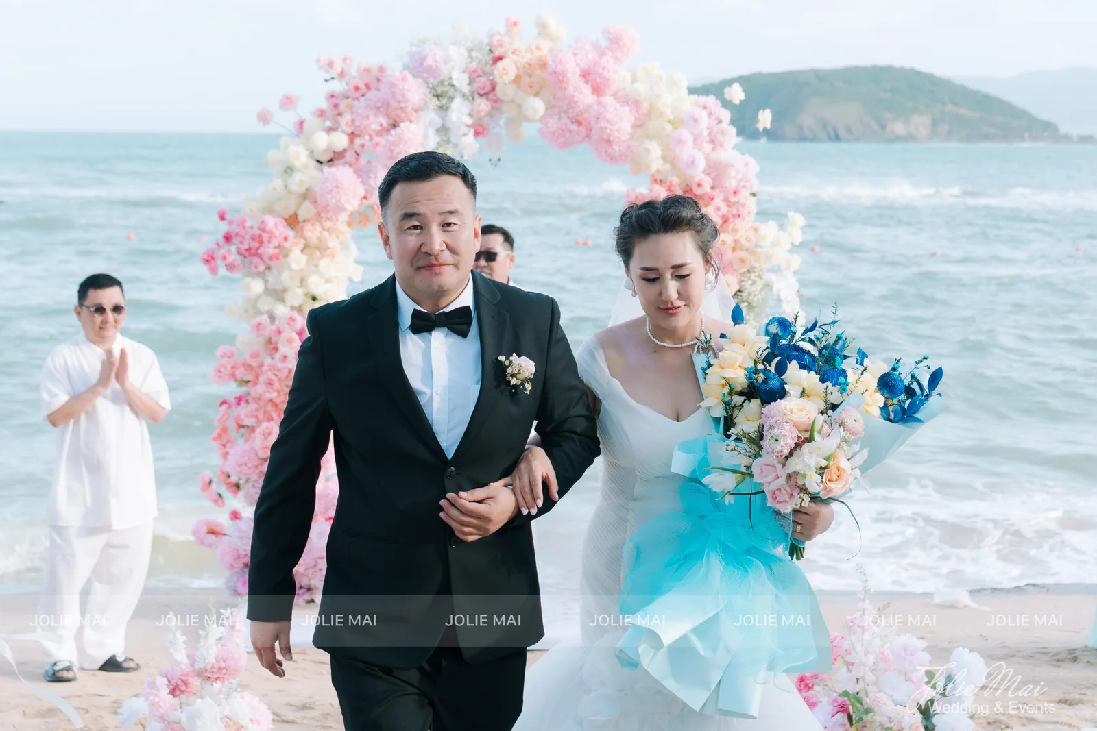 Groom and bride walking up the sand aisle, the bride holding a blue and cream bouquet tied with a turquoise ribbon Groom and bride walking up the sand aisle, the bride holding a blue and cream bouquet tied with a turquoise ribbon