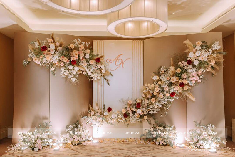 Dịch Vụ Cưới Nha Trang Một Đầu Mối: Gia Tiên, Tiệc, Mâm Quả Backdrop vách be trong sảnh khách sạn, hoa hồng kem và đỏ phối cỏ pampas chạy chéo, chữ cái đầu tên cặp đôi ở giữa