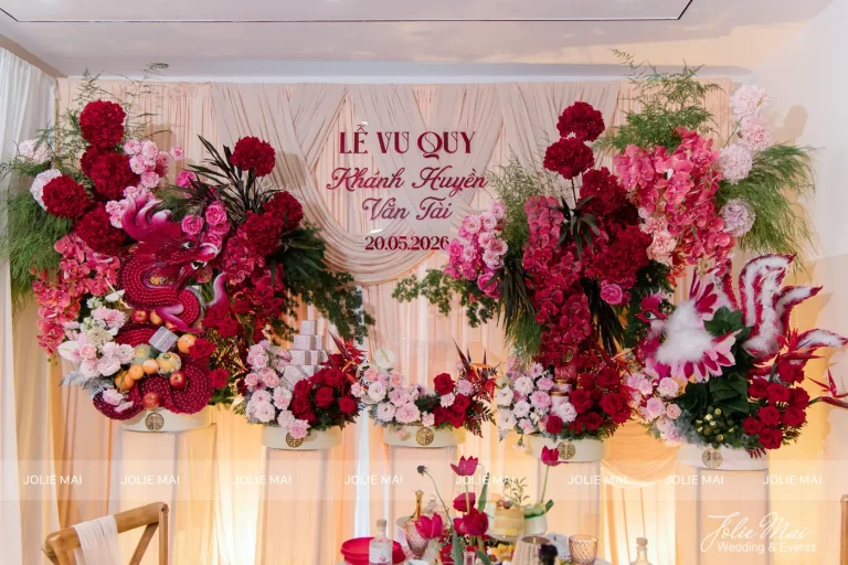 Dịch Vụ Cưới Nha Trang Một Đầu Mối: Gia Tiên, Tiệc, Mâm Quả Backdrop lễ vu quy: phông voan kem vén nếp, hai cụm hoa đỏ hồng buông tầng, tên cặp đôi ở giữa