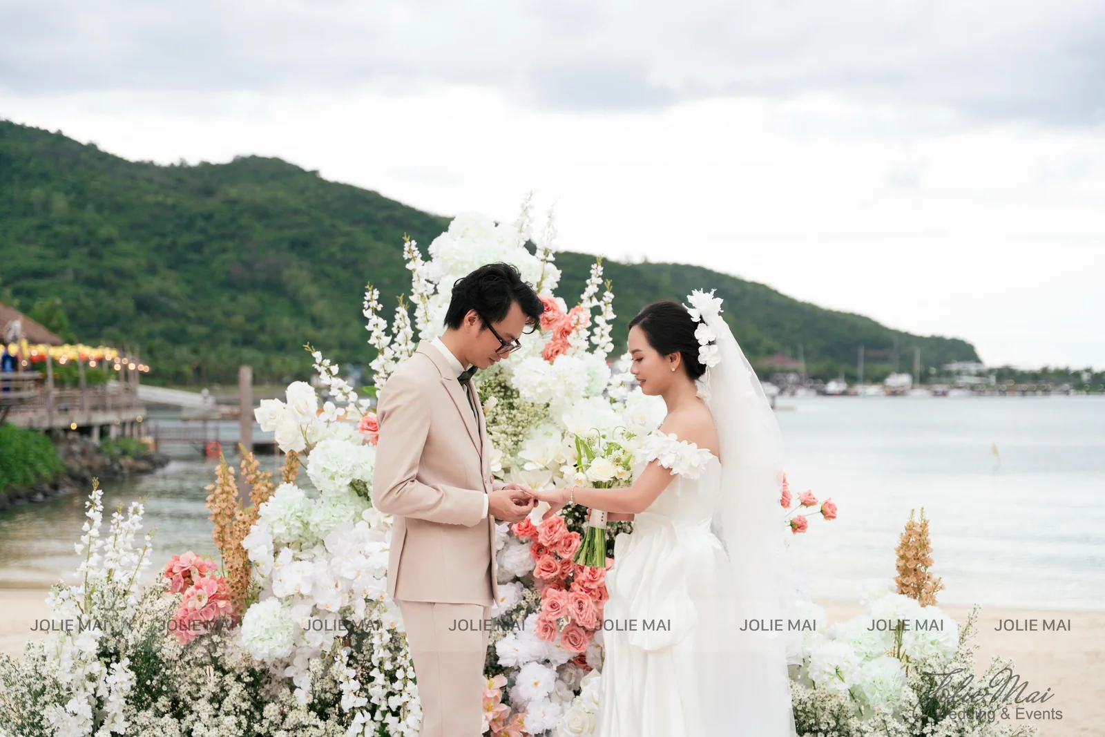 Cô dâu chú rể trao nhẫn trước cụm hoa cao trắng và cam san hô Cô dâu chú rể trao nhẫn trước cụm hoa cao trắng và cam san hô