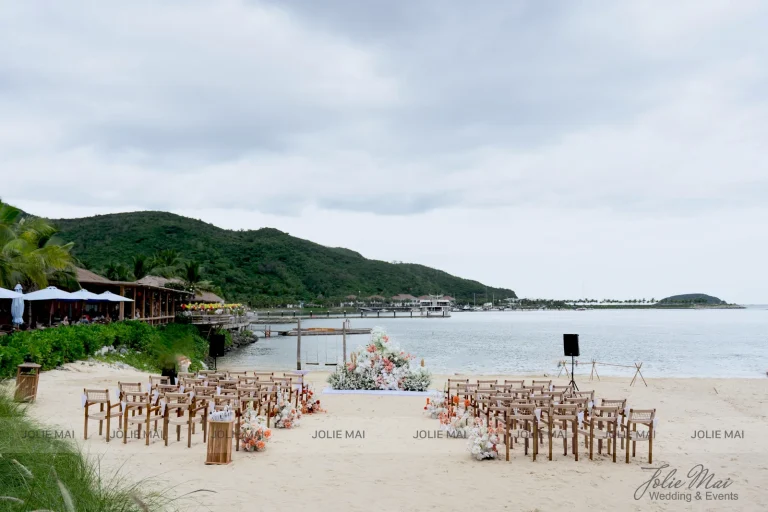 Quay Chụp Phóng Sự Cưới Nha Trang: Một Đầu Mối Từ Trang Trí Đến Thước Phim Sân lễ trên bãi cát nhìn ra vịnh Nha Trang: cụm hoa tròn trên thảm trắng, hai khối ghế gỗ đối xứng, bến gỗ và đồi xanh phía sau