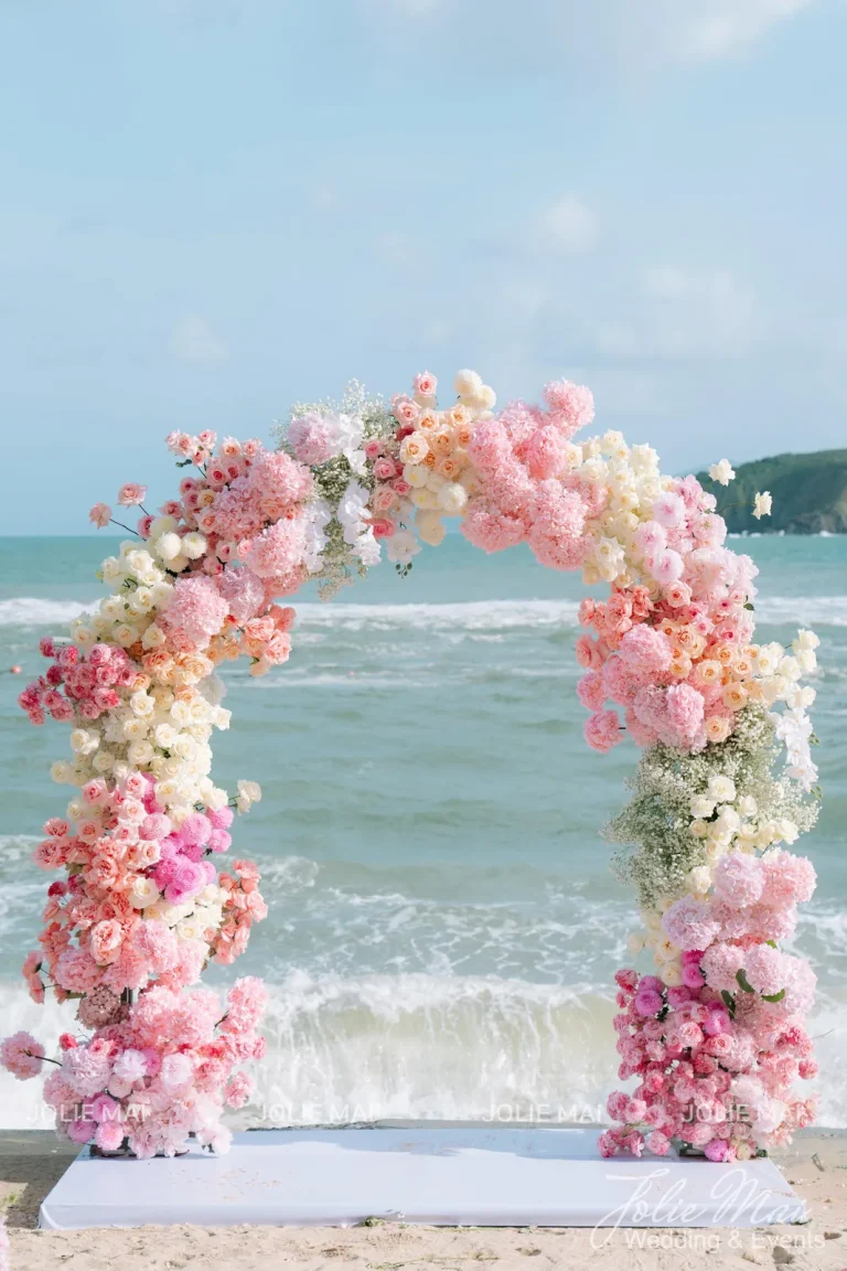Dịch Vụ Cưới Nha Trang Một Đầu Mối: Gia Tiên, Tiệc, Mâm Quả Dịch vụ cưới Nha Trang trên bãi biển: cổng vòm hoa chuyển hồng đậm, cam đào, trắng kem trên thảm trắng sát mép sóng