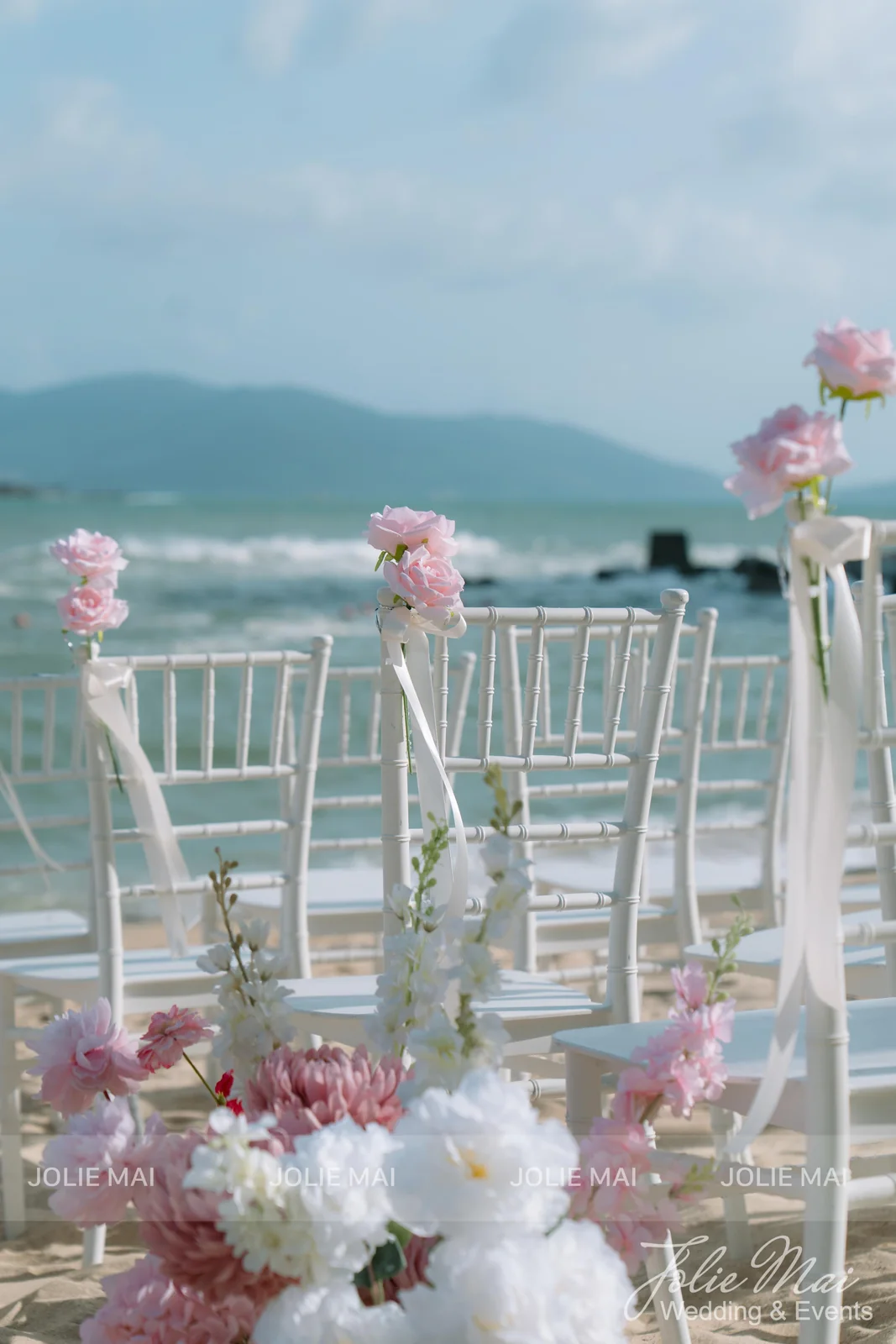 Hàng ghế tiffany trắng buộc nơ voan trên cát, đầu hàng gắn hoa hồng Hàng ghế tiffany trắng buộc nơ voan trên cát, đầu hàng gắn hoa hồng