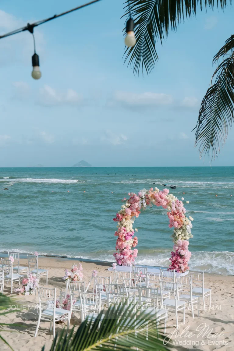 Dịch Vụ Cưới Nha Trang Một Đầu Mối: Gia Tiên, Tiệc, Mâm Quả Cổng vòm hoa hồng phấn nhìn từ xa, khung bởi dây đèn bóng treo và tán dừa, ghế trắng xếp trước cổng