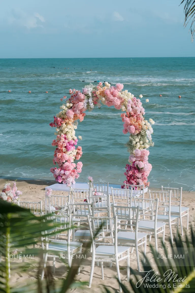 Dịch Vụ Cưới Nha Trang Một Đầu Mối: Gia Tiên, Tiệc, Mâm Quả Cổng vòm hoa hồng phấn nhìn qua lá dừa, hàng ghế Tiffany trắng trải trên cát dẫn tới cổng
