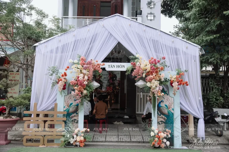 Dịch Vụ Cưới Nha Trang Một Đầu Mối: Gia Tiên, Tiệc, Mâm Quả Lều voan trắng dựng trước nhà với bảng Tân Hôn, hai cụm hoa cam hồng và chữ Song Hỷ gỗ bên trái