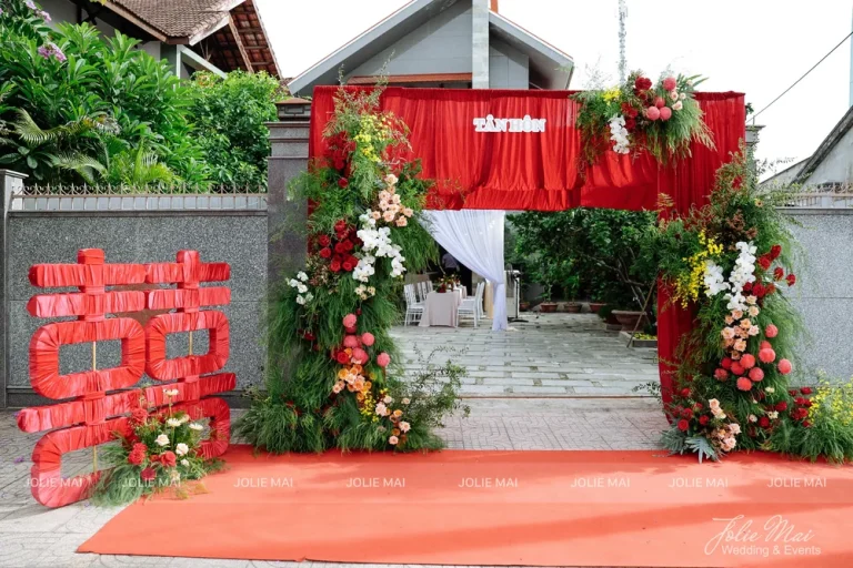 Dịch Vụ Cưới Nha Trang Một Đầu Mối: Gia Tiên, Tiệc, Mâm Quả Chính diện cổng rèm nhung đỏ giữa hai trụ hoa cao, chữ Song Hỷ đỏ bên trái và thảm cam dài