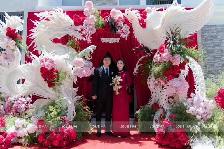 Quay Chụp Phóng Sự Cưới Nha Trang: Một Đầu Mối Từ Trang Trí Đến Thước Phim Cổng lễ gia tiên rèm nhung đỏ giữa hai tạo hình phượng trắng kết hoa hồng, hai người mặc áo đỏ đứng giữa
