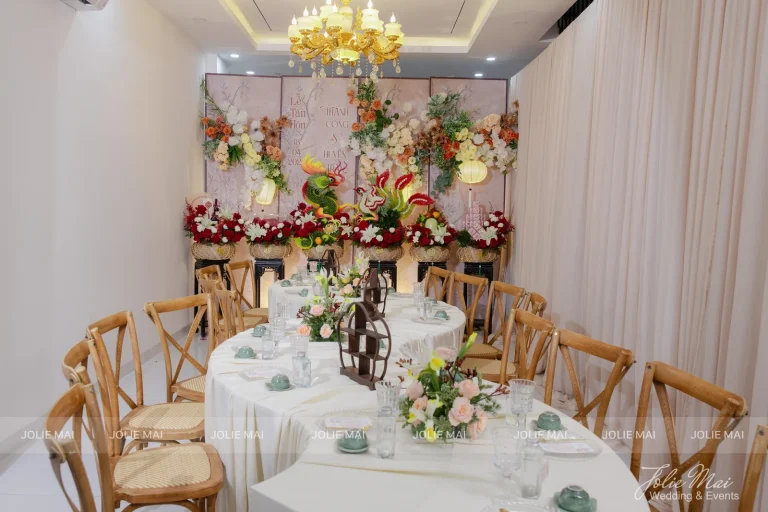 Quay Chụp Phóng Sự Cưới Nha Trang: Một Đầu Mối Từ Trang Trí Đến Thước Phim Phòng tiệc nhỏ bàn tròn khăn kem, ghế gỗ cross-back, hoa hồng đỏ cam giữa bàn, đèn chùm vàng và phông hoa cuối phòng