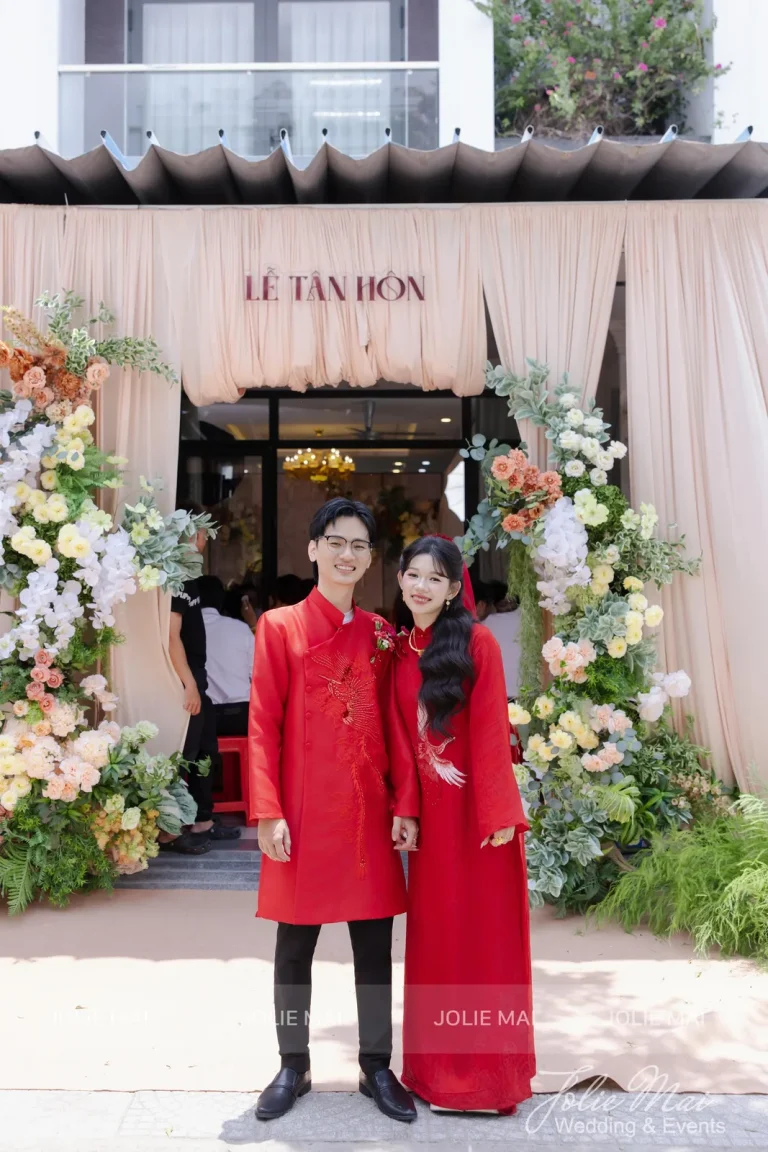 Dịch Vụ Cưới Nha Trang Một Đầu Mối: Gia Tiên, Tiệc, Mâm Quả Cô dâu chú rể áo dài đỏ đứng trước cổng hoa voan hồng đào có bảng Lễ Tân Hôn, nắng ngoài trời