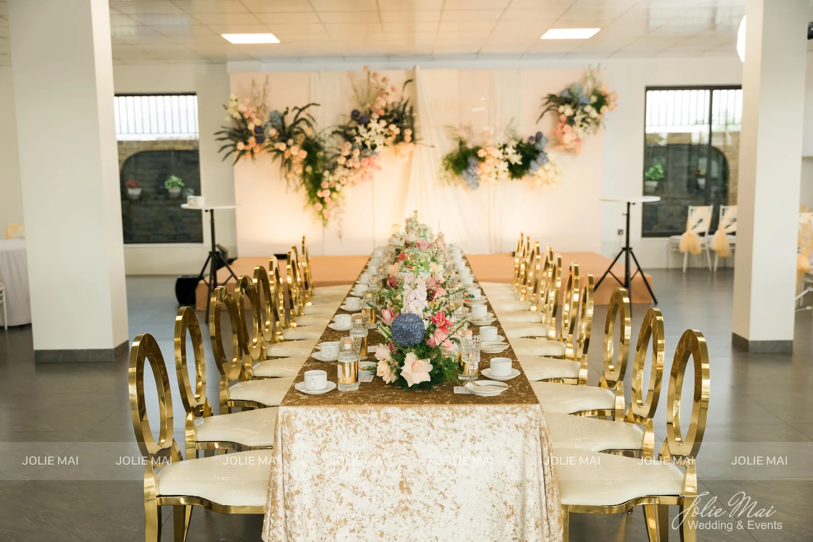 Toàn cảnh sảnh tiệc cưới với bàn dài khăn nhũ vàng, hai dãy ghế vàng và backdrop voan Toàn cảnh sảnh tiệc cưới với bàn dài khăn nhũ vàng, hai dãy ghế vàng và backdrop voan