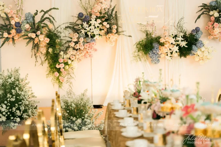 Dịch Vụ Cưới Nha Trang Một Đầu Mối: Gia Tiên, Tiệc, Mâm Quả Backdrop với cụm hoa treo từ trần xuống, bóng bay trắng ở hai bên tiền cảnh, bàn khăn nhũ vàng và ghế vàng bên phải khung