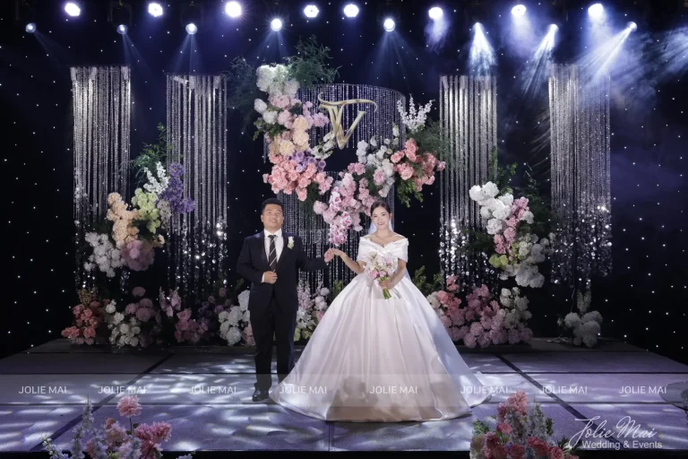 Dịch Vụ Cưới Nha Trang Một Đầu Mối: Gia Tiên, Tiệc, Mâm Quả Cô dâu chú rể trên sân khấu nền sao, bốn rèm pha lê rủ, vòm hoa hồng phấn treo giữa với chữ lồng và các cụm hoa dưới sàn kính
