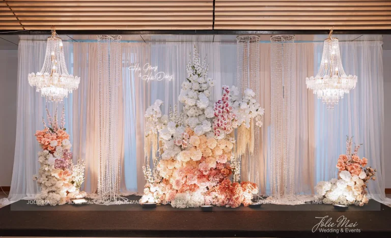 Dịch Vụ Cưới Nha Trang Một Đầu Mối: Gia Tiên, Tiệc, Mâm Quả Backdrop rèm voan kem với hai đèn chùm pha lê và khối hoa hồng, đào, trắng dựng tầng giữa phông