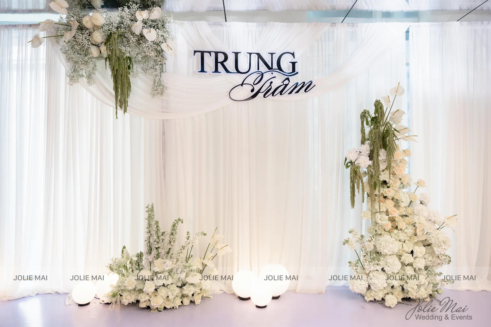 Backdrop voan trắng vắt nếp, bảng tên chữ nổi và tháp hoa trắng cao trong sảnh tiệc Backdrop voan trắng vắt nếp, bảng tên chữ nổi và tháp hoa trắng cao trong sảnh tiệc